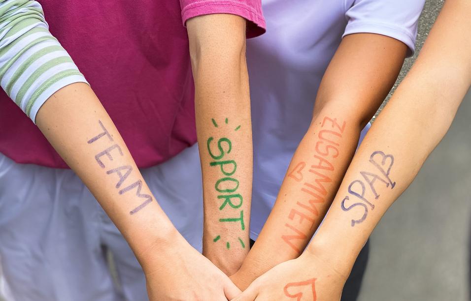 Vier Arme liegen übereinander, mit farbiger Schrift: „TEAM“, „SPORT“, „ZUSAMMEN“, „SPASS“ auf der Haut.