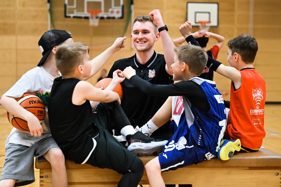 Sechs Kinder und ein Trainer zeigen Freude beim Basketballtraining auf einer Holzbank in einer Sporthalle.