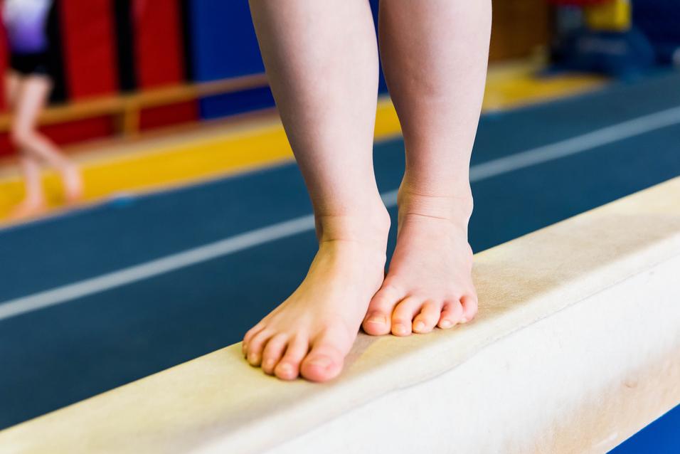 Füße eines Kindes stehen auf einem schmalen Turnbalken in einer Sporthalle.