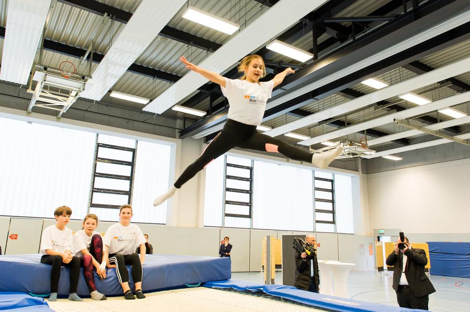 Mädchen springt mit ausgestreckten Armen über eine Trampolinfläche, während Zuschauer auf einer Bank sitzen.
