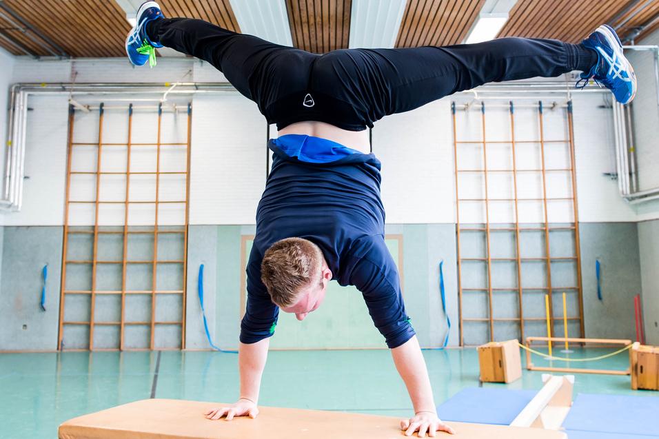 Junge macht Handstand auf einem Holztisch in einer Sporthalle mit verschiedenen Turngeräten im Hintergrund.
