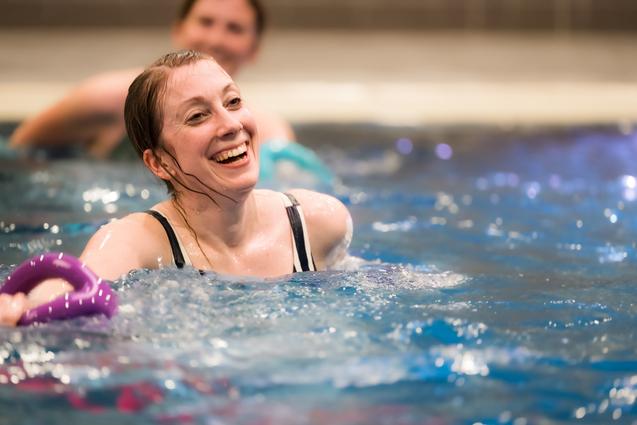Lächelnde Frau im Wasser hält einen lila Schwimmring, im Hintergrund sind weitere Schwimmer zu sehen.