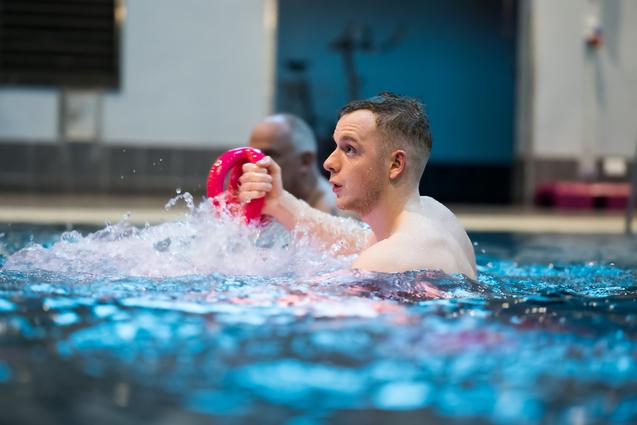 Mann im Wasser hält einen pinken Kegel und spritzt Wasser, im Hintergrund eine weitere Person.