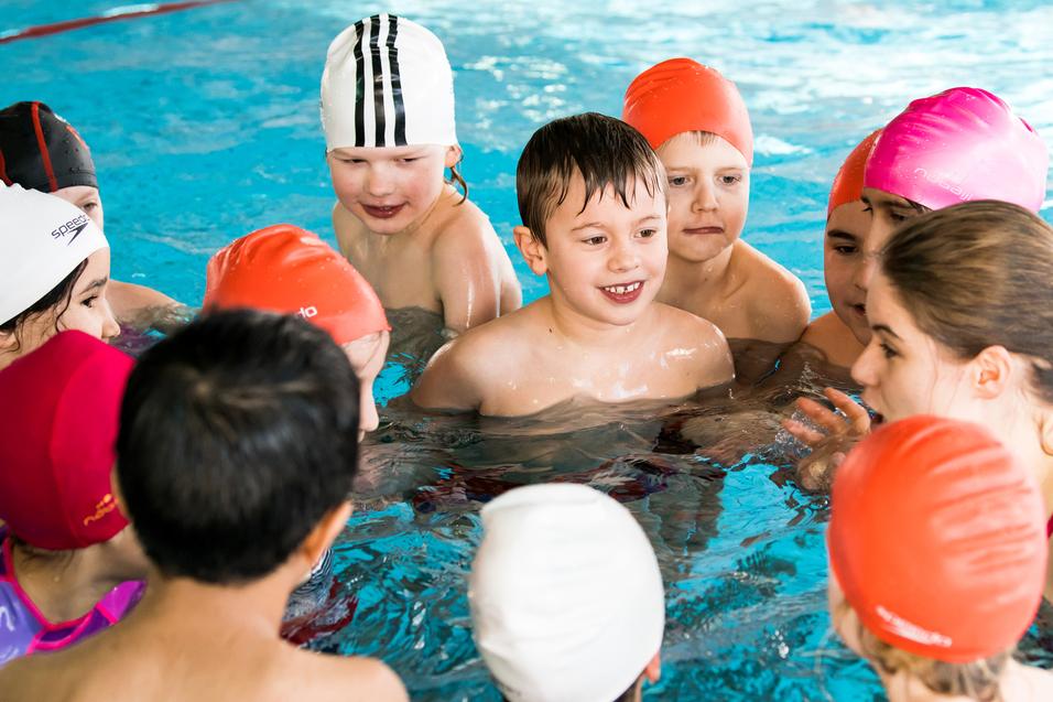 Gruppe von Kindern im Schwimmbecken, die sich um eine Trainerin versammeln und miteinander kommunizieren.